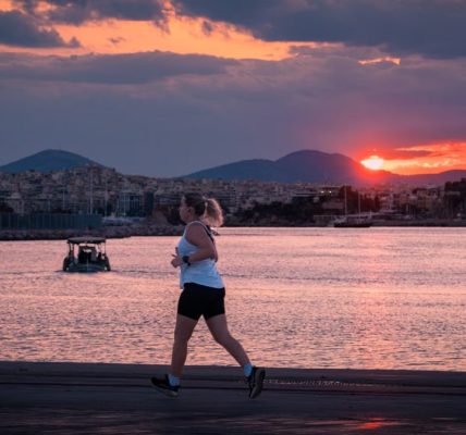 A woman jogs along the waterfront at sunset with a cityscape and boat in the background.