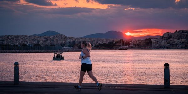 A woman jogs along the waterfront at sunset with a cityscape and boat in the background.